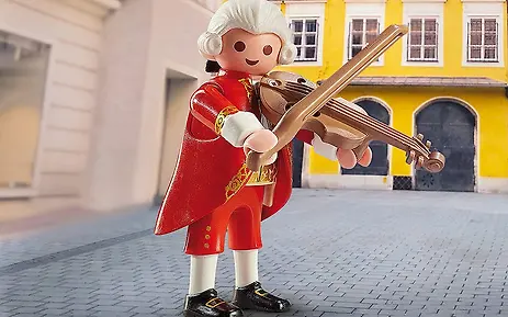Playmobil-Figur in roter historischer Kleidung mit weißer Perücke, die eine Violine vor einem gelben Gebäude mit Arkaden hält.