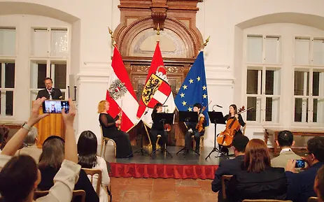 Classical hall with music ensemble and Austrian flags during a ceremonial event at the University of Salzburg.