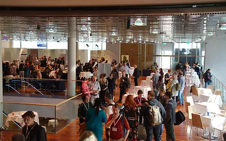 Conference or event space with modern architecture featuring numerous visitors at white standing tables and furniture.