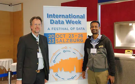 Two men stand at the International Data Week 2023 in Salzburg in front of an event poster, one in traditional attire, the other in casual clothing.