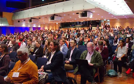 Large conference hall with fully occupied rows and modern lighting. Numerous participants attending an event in a professional setting.