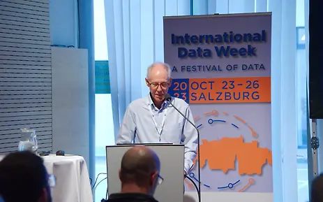Scholar presenting at the International Data Week conference in Salzburg, standing at a podium with event banner.