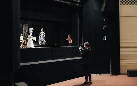 In der dunklen Theaterbühne stehen mehrere stilisierte Puppen in prächtigen historischen Gewändern - eine weiße Dame, eine silberne Figur und ein Herr im roten Rock. Eine Person im Vordergrund dokumentiert diese surreale Szenerie mit einer Kamera, während die Bühne eine geheimnisvolle und theatralische Atmosphäre ausstrahlt. Theaterbühne mit historischen Puppen in Kostümen, eine Person fotografiert die szenische Anordnung von verschiedenen Figuren.
