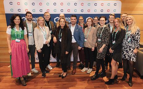 Eine bunte Delegation versammelt sich beim Europe Congress, wo Professionalität auf Individualität trifft. Die Teilnehmenden stehen entspannt nebeneinander, in einem Raum mit warmen Holzböden und moderner Wandgestaltung. Ihre Kleidung reicht voneleganten Businessanzügen bis hin zu einem traditionellen Dirndl und vermittelt eine offene, internationale Atmosphäre. Gruppenaufnahme von etwa 15 Personen bei einer Konferenz mit blau-rotem Hintergrund und Firmenlogo, unterschiedlich gekleidet in professioneller Businesskleidung.