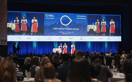 In einem stilvollen Konferenzsaal in Salzburg präsentieren drei Frauen in eleganter Tracht einen kulturellen Moment während des internationalen MCE-Kongresses. Die blaue Bühnenbelechtung und der volle Saal zeugen von einer professionellen und inspirierenden Veranstaltung im Herzen Österreichs. Drei Frauen in traditioneller rot-blauer Tracht auf der Bühne einer Konferenz des MCE Central & Eastern Europe Kongresses in Salzburg.