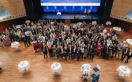 In einem lichtdurchfluteten Konferenzsaal versammeln sich zahlreiche Teilnehmer zu einem Gruppenfoto. Die Holzdielen des Bodens und die blau schimmernde Bühnendekoration verleihen dem Raum eine professionelle und gleichzeitig einladende Atmosphäre. Weiß gedeckte Stehtische verteilen sich im Vordergrund und rahmen die Szenerie stilvoll ein. Große Gruppenaufnahme bei einer Konferenz mit vielen Teilnehmenden in einem modernen Konferenzsaal mit Holzboden und blauer Bühnendekoration.