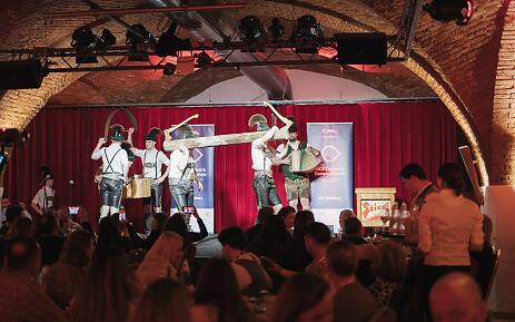 In einem stimmungsvoll beleuchteten Backsteingewölbe präsentiert eine Folkloregruppe in traditioneller Lederhosen-Tracht einen lebendigen Auftritt. Die rote Bühnenvorhang und die Konzertbeleuchtung schaffen eine atmosphärische Kulisse für dieses kulturelle Erlebnis im Herzen Salzburgs. Traditionelle Folkloregruppe auf einer Bühne mit Ziegelgewölbe, die bayerische oder österreichische Trachtenkultur in einer intimen Veranstaltung präsentiert.