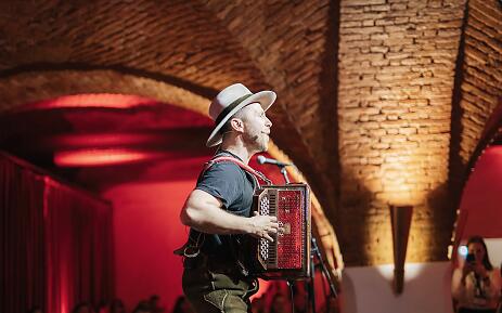 In einem stimmungsvollen Backsteingewölbe entfaltet sich ein musikalischer Moment: Ein Akkordeonist in elegantem weißem Hut steht im warmen Rot der Bühnenbeleuchtung. Seine Körperhaltung verrät Leidenschaft und Hingabe an die Musik, während er sein rotes Akkordeon virtuos spielt. Akkordeonist in weißem Hut auf einer Bühne mit ziegelsteinernen Wänden und roter Beleuchtung, konzentriert beim Musizieren.