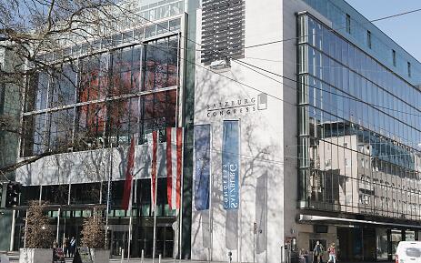 Der Salzburg Congress präsentiert sich als architektonisches Highlight mit transparenten Glasfassaden und klaren Linien. Elegante Flaggen rahmen den Eingang ein, während winterliche Bäume sanfte Schatten auf die moderne Architektur werfen. Ein repräsentativer Ort für Kongresse und Veranstaltungen im Herzen von Salzburg. Moderner Glasfassaden-Komplex des Salzburg Congress mit österreichischen und europäischen Flaggen im Vordergrund, umgeben von kahlen Winterbäumen.
