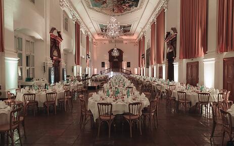 Ein majestätischer Saal erstrahlt in elegantem Glanz, mit kunstvoll verzierten Decken, prächtigen Kronleuchtern und edlen roten Vorhängen. Die sorgfältig gedeckten weißen Tische laden zu einem exklusiven Ereignis ein, während das barocke Ambiente eine zeitlose Atmosphäre von Grandezza und Perfektion ausstrahlt. Festlicher Veranstaltungssaal mit barocker Architektur, weiß-roten Vorhängen und eleganten runden Tischen für ein Bankett oder Gala-Dinner.