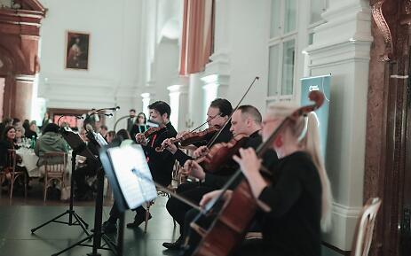 In einem prachtvollen Saal mit hohen Decken und klassizistischer Architektur entfaltet ein Streichquartett seine musikalische Kunst. Die Musiker in schwarzer Konzertkleidung spielen konzentriert ihre Instrumente, während im Hintergrund Zuhörer an gedeckten Tischen sitzen und der Musik lauschen. Streichquartett bei einem Konzert in einem eleganten, historischen Saal mit weißen Säulen und gedämpfter Beleuchtung.