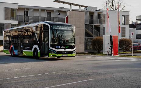 Moderner elektrischer Stadtbus in grün-weißer Farbgebung an einer Ladestation vor zeitgenössischer Architektur.