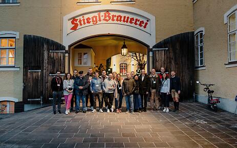 In warmem Licht erstrahlt der legendäre Eingang der Stiegl-Brauerei, wo eine bunte Gruppe von Menschen eng beieinander steht. Die historische Backsteinmauer und die alten Holztore erzählen von jahrhundertealter Brautradition und österreichischer Gastlichkeit. Gruppenaufnahme vor dem historischen Eingang der Stiegl-Brauerei mit rustikaler Backsteinarchitektur und Holztoren in Salzburg.