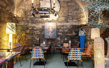 An atmospheric dining room with a rustic stone wall, colorful designer chairs, and a Mediterranean flair. Artistic lighting and a central portrait lend the space a creative, warm character with a southern spirit. Colorful dining room with stone wall, art portrait, designer chairs, and Mediterranean décor.