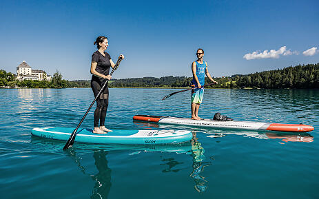 Amidst a breathtaking lake landscape, two paddlers glide on their stand-up paddleboards through crystal-clear water. The turquoise water surface reflects the brilliant blue sky, while a majestic historic building and dense forests frame the scenery in the background. Two people paddle on turquoise stand-up paddleboards on a clear lake with forested shores and a historic building in the background.