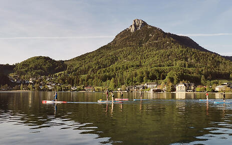 Amidst a breathtaking mountain landscape, paddleboarders glide gently across the mirror-smooth water surface. A mighty rocky peak towers over an idyllic village, while firs and deciduous trees bathe the slopes in lush green. The tranquility and vastness of the landscape invite you to linger and take a deep breath. Paddleboarders on a calm mountain lake with green forests and a distinctive rocky mountain in the background, surrounded by a picturesque village.