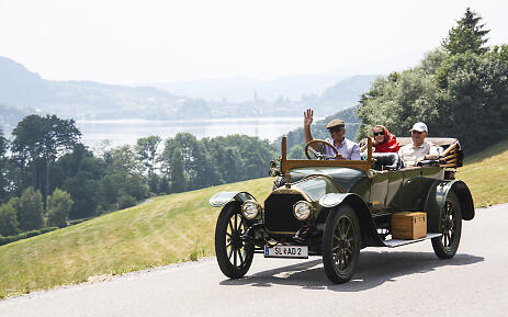 A nostalgic convertible from the early days of automobiles glides smoothly along a winding mountain road. The passengers enjoy the ride in summer attire, while a breathtaking alpine landscape with a blue lake and lush green slopes unfolds in the background. The scenery breathes pure joy of life and the spirit of exploration from bygone times. Vintage convertible car driving on a mountain road with a scenic lake and forested mountain slopes in the background.