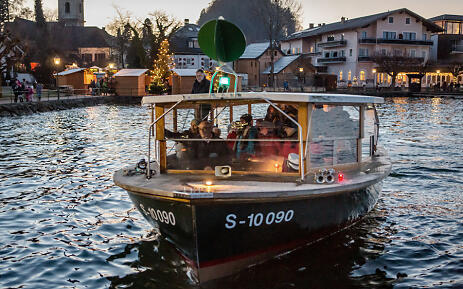 In the twilight, a cozy excursion boat glides across the calm water. Christmas-lit market stalls and festively decorated houses line the shore, while the boat with its passengers radiates a warm, inviting atmosphere. Illuminated excursion boat on a winter waterway with Christmas market and historic village backdrop.