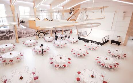 At fahr(T)raum, technological history and modern event culture merge into an impressive total work of art. A historic aircraft hovers above festively set tables, while the light-flooded ambiance radiates timeless elegance. The white and red color composition and architectural precision make this space an extraordinary venue. Elegant event space with vintage biplane, white-covered tables and red chairs in a bright, airy building.