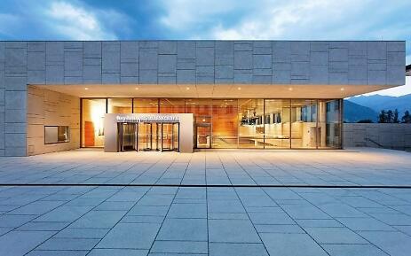 The Ferry Porsche Congress Center presents itself as an architectural masterpiece with clear lines and elegant light staging. The geometric facade of gray concrete panels harmoniously contrasts with the warm wooden tones of the entrance hall and the expansive glass fronts. Soft mountain contours are silhouetted against the blue evening sky in the background. Modern congress center with gray facade and large glass fronts, warmly illuminated at evening with mountain panorama in background.