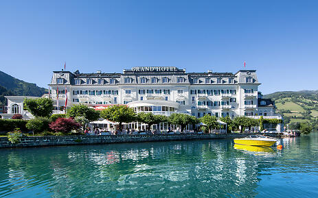 The Grand Hotel in Zell am See stands majestically on the shore of the crystal-clear lake, embedded in the breathtaking mountain landscape of Salzburg. The white, historic hotel architecture is reflected in the emerald green waters, while a yellow yacht in the foreground completes the idyllic scene. Luxurious Grand Hotel situated directly on the turquoise lake shore, surrounded by green mountains and blue sky.