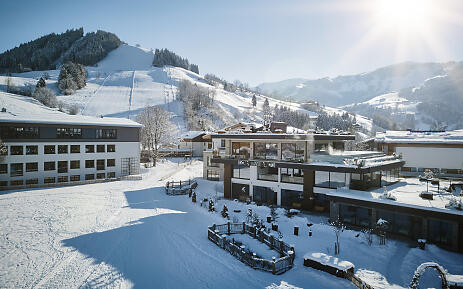 Die Winterlandschaft der Hochkönigin präsentiert sich in atemberaubender Pracht: Schneeweiße Skipisten schlängeln sich sanft die Berghänge hinab, umrahmt von dicht verschneiten Tannenwaldhängen. Moderne, kubische Architektur fügt sich harmonisch in die alpine Szenerie ein, mit Gebäuden, die transparente Glasfassaden und geradlinige Formen aufweisen und einen zeitgemäßen Kontrast zur ursprünglichen Bergwelt bilden. Winterliche Berglandschaft mit modernen Gebäuden und verschneiten Skipisten im Salzburger Land, umgeben von bewaldeten Berghängen und strahlend blauem Himmel.