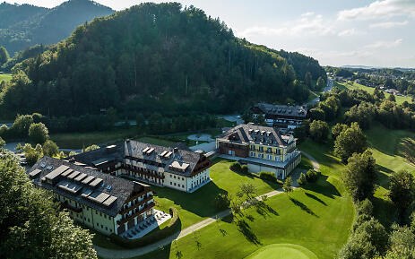 Aerial view of a luxurious resort with multiple buildings nestled in a green mountain landscape with meadows and forests.