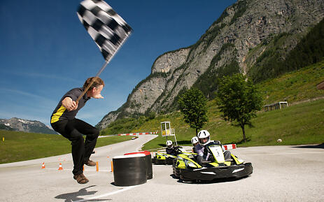 Inmitten einer atemberaubenden Berglandschaft des Salzburger Landes entfaltet sich eine Go-Kart-Strecke, die Adrenalin und Naturschönheit perfekt verbindet. Ein Rennleiter mit karierter Flagge dirigiert die Fahrer auf der sonnendurchfluteten Strecke, während imposante Felswände den Horizont säumen und grüne Wiesen den Rand der Rennstrecke schmücken. Go-Kart-Strecke vor alpiner Bergkulisse mit Rennleiter, der eine karierte Flagge schwenkt und Fahrern in Rennanzügen.