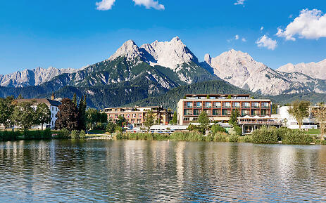 Nestled in the breathtaking mountain landscape of Salzburg, the Hotel Ritzenhof sits like an architectural gem. Modern hotel buildings harmoniously blend into the lakeshore, while imposing Alpine rock peaks form a dramatic backdrop. The clear water surface reflects the architecture and surrounding natural scenery in gentle wave movements. Modern hotel by a lake shore with majestic Alpine mountains in the background, surrounded by lush vegetation and reflective water surface.