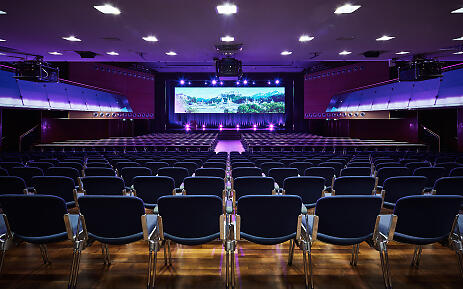 Moderner Konferenz- und Veranstaltungssaal mit blau-violetter Beleuchtung und großer Leinwand, bestuhlt mit schwarzen Stühlen.