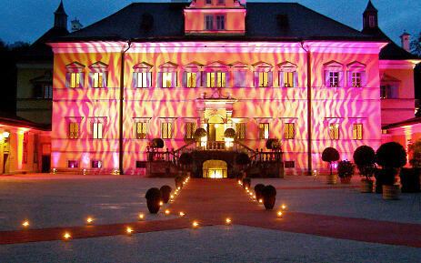 In magischem Licht erstrahlt das barocke Schloss Hellbrunn in einem spektakulären Farbenspiel aus Pink und Orange. Sanfte Kerzenlichter führen den Blick über den nächtlichen Vorplatz zur illuminierten Fassade, die wie ein leuchtender Kunstwerk in der Dunkelheit schwebt. Schloss Hellbrunn in pinken und roten Lichteffekten bei Nacht, illuminiert mit Kerzenlichtern auf dem Vorplatz und dynamischen Lichtprojektionen an der Fassade.