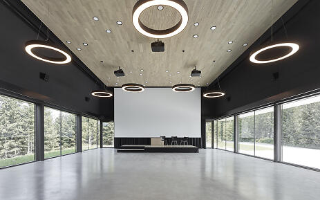 Ein architektonisch beeindruckender Raum, der Natur und Design harmonisch verschmilzt. Große Glasfenster öffnen den Blick in einen dichten Waldbestand, während die kreisförmigen Deckenleuchten eine zeitgenössische Atmosphäre schaffen. Der geräumige Raum mit betoniertem Boden und hölzerner Deckenkonstruktion wirkt gleichzeitig minimalistisch und einladend. Moderner, lichtdurchfluteter Veranstaltungsraum mit großen Glasfronten, hölzerner Decke und kreisförmigen Deckenleuchten, umgeben von Waldlandschaft.
