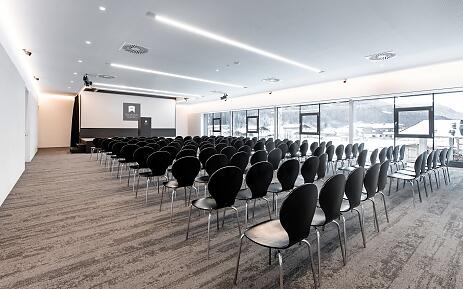 Moderner Konferenzraum mit schwarzen Stühlen, großen Glasfenstern und Bergpanorama im Hintergrund, ausgestattet für Präsentationen und Veranstaltungen.