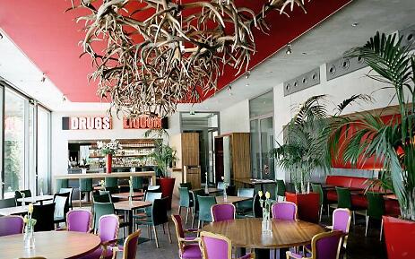 Modern restaurant M32 in the Museum der Moderne Salzburg with colorful designer chairs, eye-catching antler chandelier on the red ceiling, large windows with natural light and a view of the bar with the illuminated lettering “Drugs” and “Liquor”. Restaurant M32 with seating for guests - suitable for meals, meetings or events.