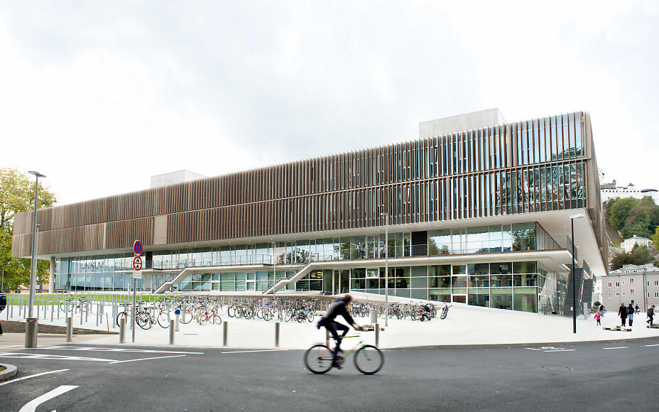 Modern building with glass facade and bicycle racks, with a cyclist in the foreground on an urban street.