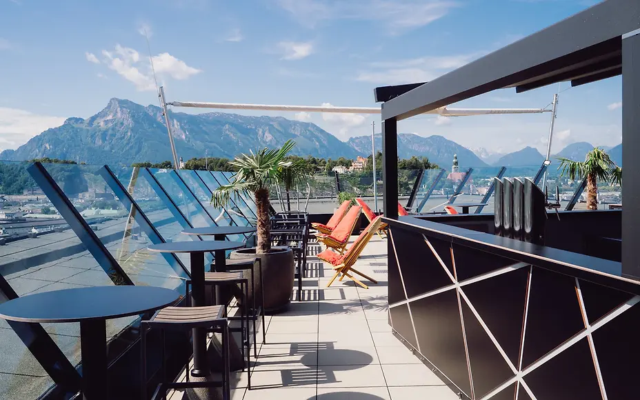 At the Arte Hotel’s rooftop bar, lounge seating, palm trees, and mountain views invite guests to relax. A highlight above Salzburg’s rooftops.