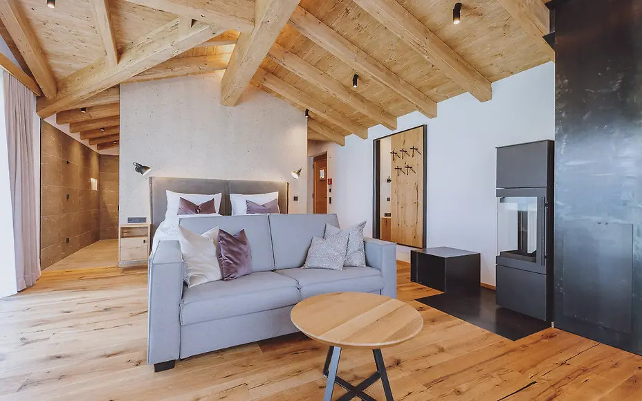 Modernes Hotelzimmer im Bergloft-Stil mit Holzdeckenbalken, grauer Couch und minimalistischer Einrichtung in warmen Holztönen.