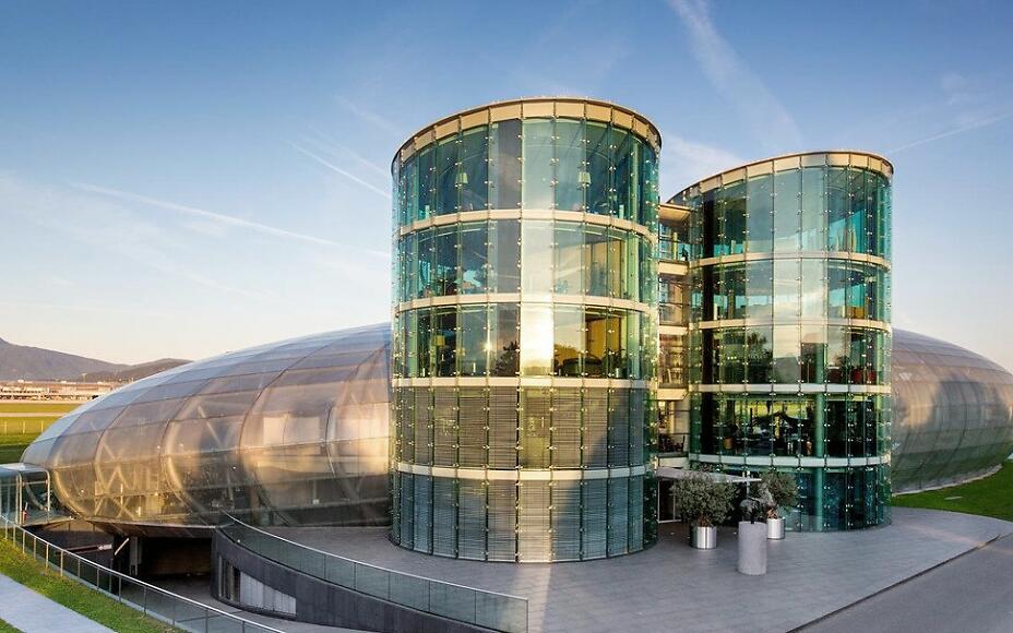 Moderner Glasarchitektur-Komplex mit zylindrischen Türmen und gewölbtem Dach, eingebettet in eine Landschaft mit Bergen im Hintergrund.