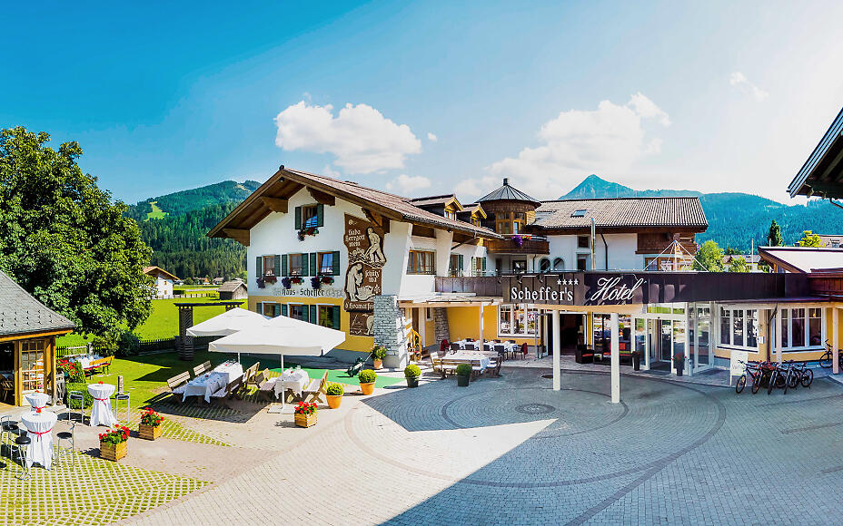 Sunlit hotel complex of Scheffer's in Altenmarkt with typical alpine architecture, surrounded by green trees and mountains in the background.