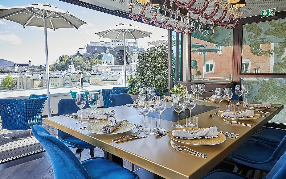 Gedeckter Restauranttisch mit Panoramablick auf die Salzburger Altstadt und Festung, blaue Samtsessel und elegante Tischdekoration.