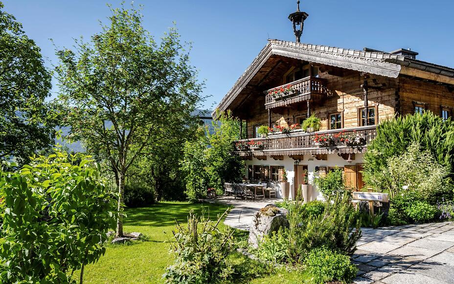 Traditional wooden chalet with wooden balcony and red flowers in a green garden, surrounded by trees and shrubs against a blue sky.