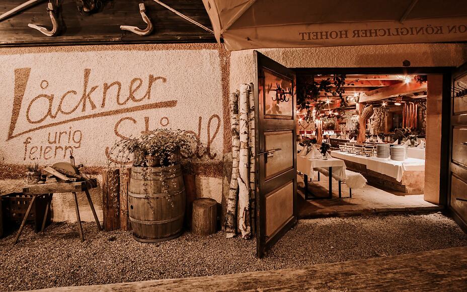 Der Eingang zum Lackner Stadl versprüht ländlichen Charme mit rustikalen Details und dem markanten Schriftzug "urig feiern". Durch die geöffnete Tür gibt der Blick das festlich gedeckte Innere frei.