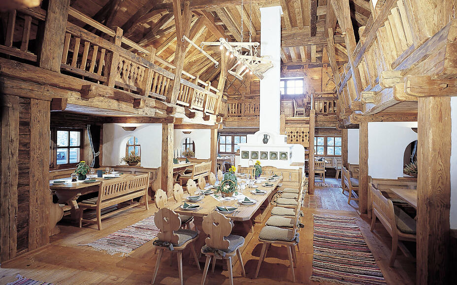 Rustic wooden interior with traditional alpine cabin architecture, featuring a large wooden table, benches, and artfully carved chairs in warm wood tones.