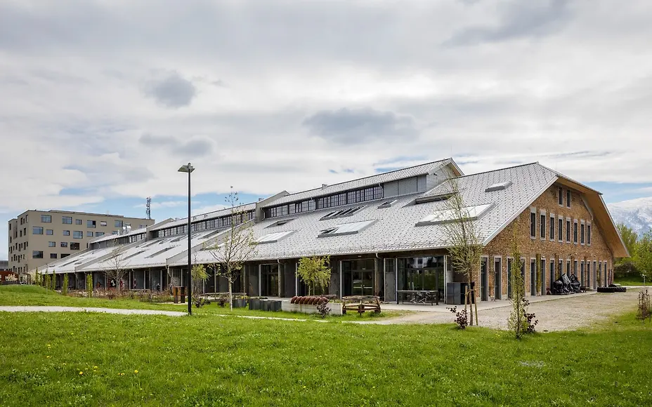 The Panzerhalle impresses with its industrial charm: A modern gabled building with large windows surrounded by greenery near the Alps.