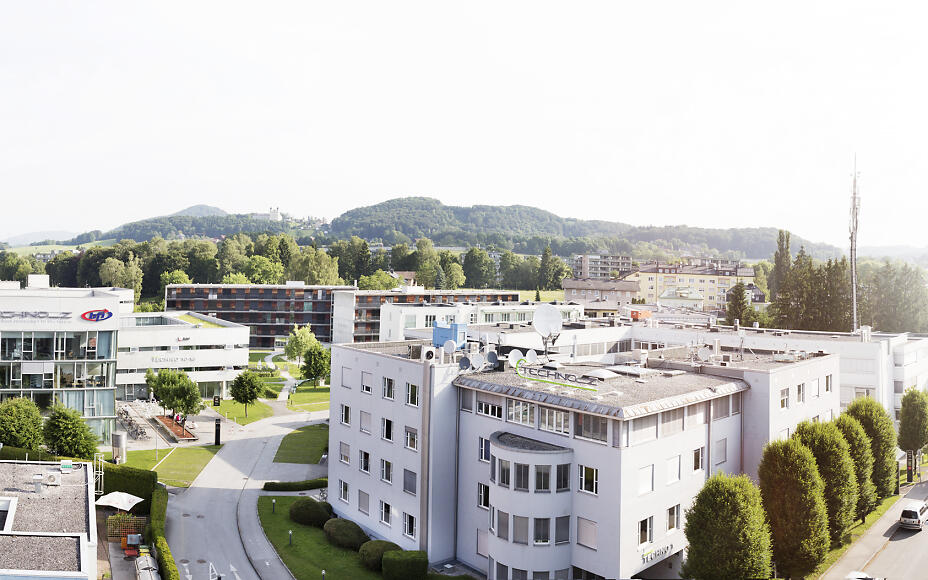 Panoramablick auf ein modernes Stadtquartier mit mehrstöckigen Gebäuden, umgeben von grünen Bäumen und bewaldeten Hügeln im Hintergrund.