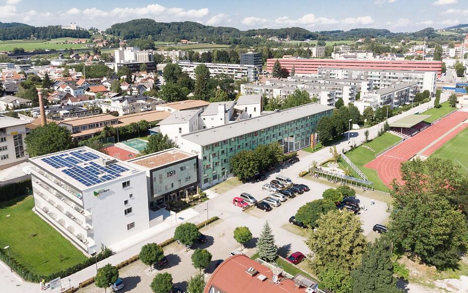 Aerial view of a modern urban landscape with building complex, parking lots, running track, and green surroundings with mountain backdrop.