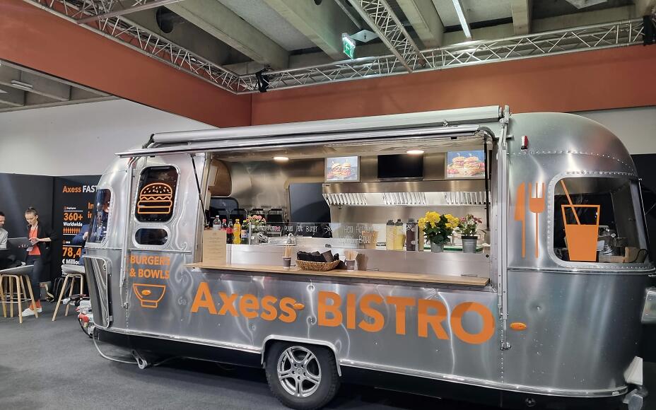Silver food truck of Axess Bistro with orange lettering, equipped for burgers and bowls, located in an exhibition hall.