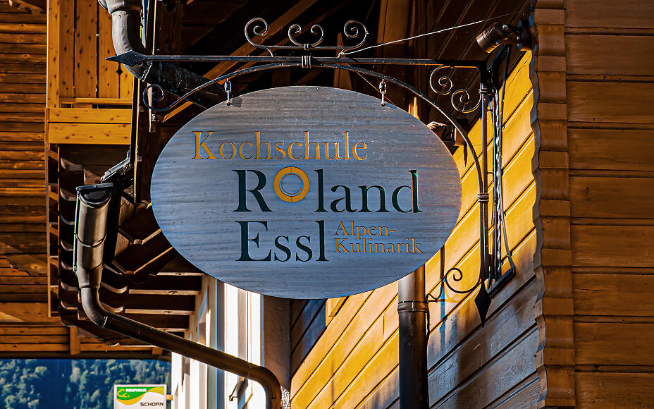 Ovales Firmenschild der Kochschule Roland Essl mit Alpen-Kulinarik-Schriftzug, angebracht an einer holzvertäfelten Wand mit warmem Lichteinfall.
