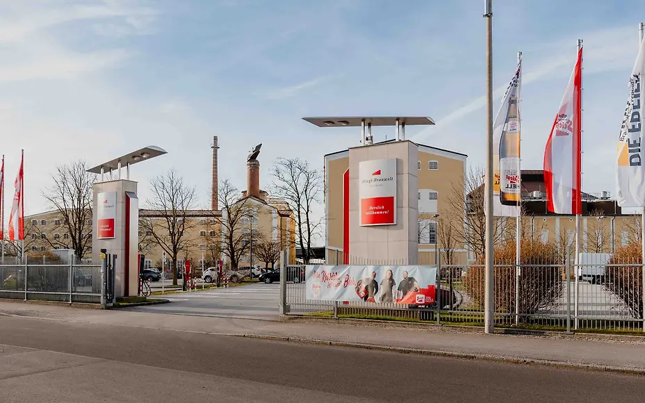 Der Eingang eines großen Brauereigeländes präsentiert sich mit Fahnen, modernen Toranlagen und historischen Gebäuden. Die Kombination aus Industriearchitektur und Markeninszenierung macht deutlich, dass hier Tradition und moderne Bierproduktion zusammenfinden.