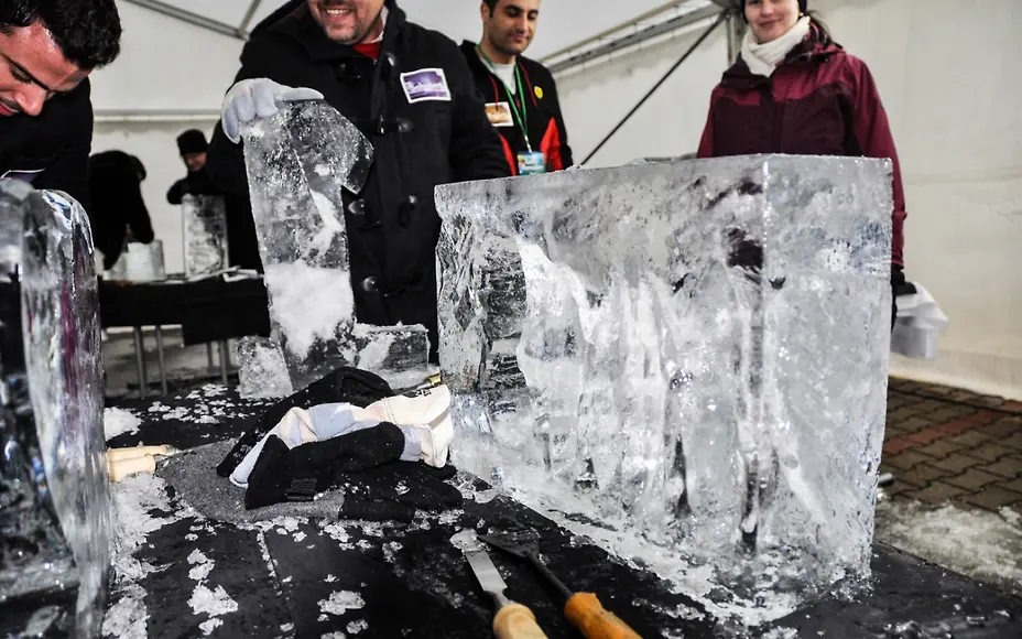 In einem geschützten Zelt arbeiten Teilnehmer konzentriert an großen Eisblöcken und formen daraus beeindruckende Skulpturen. Mit Handschuhen, Meißeln und viel Geschick entstehen aus dem transparenten Material faszinierende Strukturen, während neugierige Zuschauer den kreativen Prozess verfolgen.